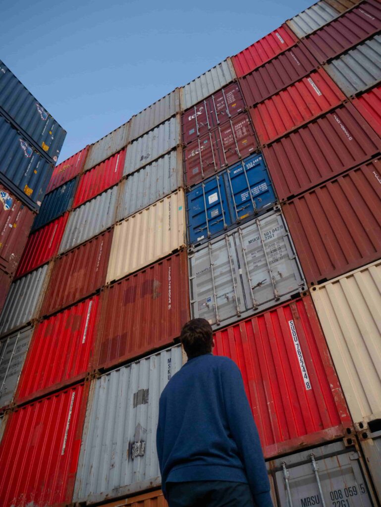 A foto mostra containers para importação, de de várias cores empilhados um sobre os outros. Na parte inferior da fotografia, um homem vestido de camisa azul de manga longa e calça também azul, inclina a cabeça para o alto e contempla a altura da estrutura montada.
